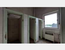 Abgenutzter Duschraum mit drei offenen Duschkabinen, weißen Wandfliesen und blassgrünen Türrahmen. Rechts befindet sich ein Fenster mit Blick auf eine Industrielandschaft sowie ein alter Heizkörper unterhalb des Fensters.