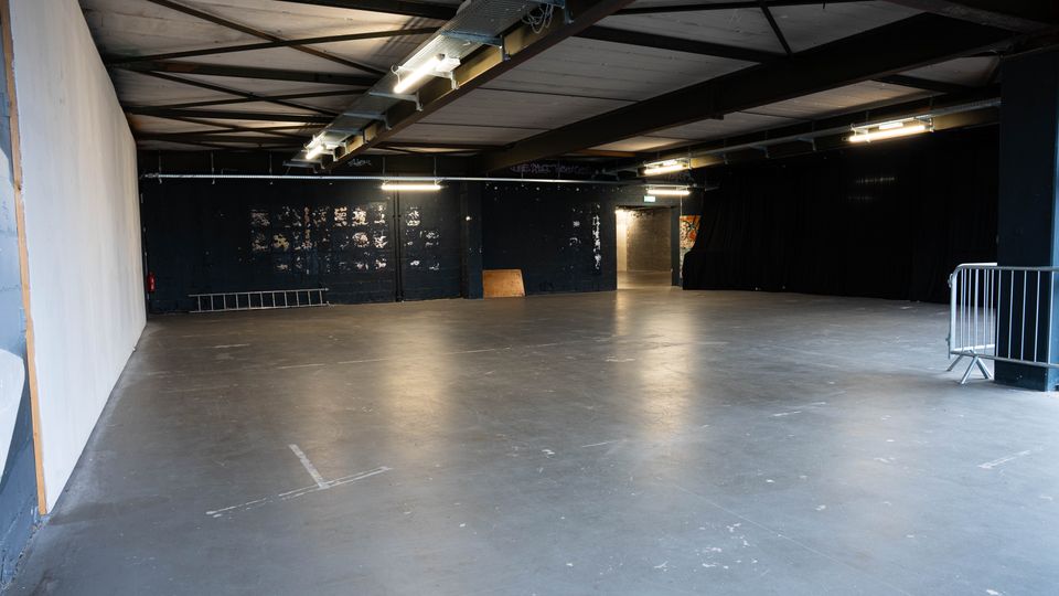 Große, leere Industriehalle mit glattem Betonboden, schwarzen Wänden und freiliegenden Stahlträgern unter der Decke. Mehrere Neonröhren und Kabeltrassen hängen an der Decke; rechts sind schwarze Vorhänge und ein Absperrgitter, hinten ein Durchgang zu einem weiteren Bereich.