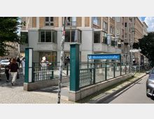Eingang zur U-Bahn-Station Weinmeisterstraße in Berlin, umgeben von einem Wohn- und Geschäftshaus mit verzierten Fassadenelementen. Im Vordergrund sind Passanten, Fahrräder sowie angrenzende Straßenschilder und parkende Autos sichtbar.
