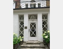 Weiße Hausfassade mit einer weißen, verglasten Eingangstür und dekorativen Gittern in einem geschwungenen Muster, flankiert von großen Fenstern mit identischen Ornamenten. Vor der Tür führen Steinstufen hinauf, eingerahmt von blühenden Hortensien in Korbkübeln.