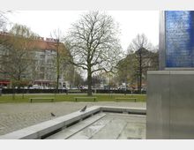 Ein Platz mit gepflasterten Wegen, Sitzbänken und einer Rasenfläche, umgeben von Bäumen; im Hintergrund befindet sich ein mehrstöckiges Gebäude mit rotem Dach und Schriftzug "Block House". Im Vordergrund steht ein modernes Denkmal mit einer blauen Glasplatte, auf der Schrift zu erkennen ist.