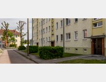 Mehrstöckiges Wohngebäude mit hellgelber Fassade und rechteckigen weißen Fenstern, flankiert von kleinen Grünflächen und Hecken. Im Hintergrund steht ein weiteres Gebäude mit orangefarbener Fassade und großen Glasfenstern, umgeben von Bäumen.