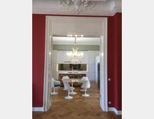 Blick durch eine Tür mit einer reich verzierten weißen Stuckdecke auf eine moderne Küche mit weißen Hochglanzmöbeln, einem runden Esstisch und weißen, tulpenförmigen Stühlen. Der Raum hat rote Wände und einen Boden aus hellem Fischgrätparkett.