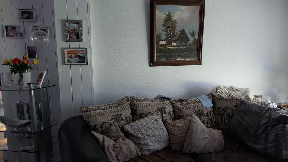 Gemütliches Wohnzimmer mit dunklem Sofa und vielen Kissen vor einer weißen Wand, darüber ein gerahmtes Landschaftsgemälde. Links steht ein gläsernes Eckregal mit Vase und Blumen vor einer vertäfelten Wand mit mehreren gerahmten Fotos.