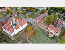 Luftaufnahme eines historischen Komplexes mit weißen, rotgedeckten Gebäuden, darunter ein Schloss mit markantem Turm, eine Kirche mit spitzem Turm und weitere Klosterbauten. Umgeben von Grünflächen, Bäumen und kleinen Straßen in einer ländlichen Umgebung.