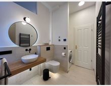 Modernes Badezimmer mit hellen Fliesen an Wand und Boden. Zu sehen sind ein Rundspiegel mit Beleuchtung, ein Holzwaschtisch mit weißem Aufsatzwaschbecken, ein wandhängendes WC, ein schwarzer Handtuchheizkörper sowie eine Tür, hinter der eine Waschmaschine steht.