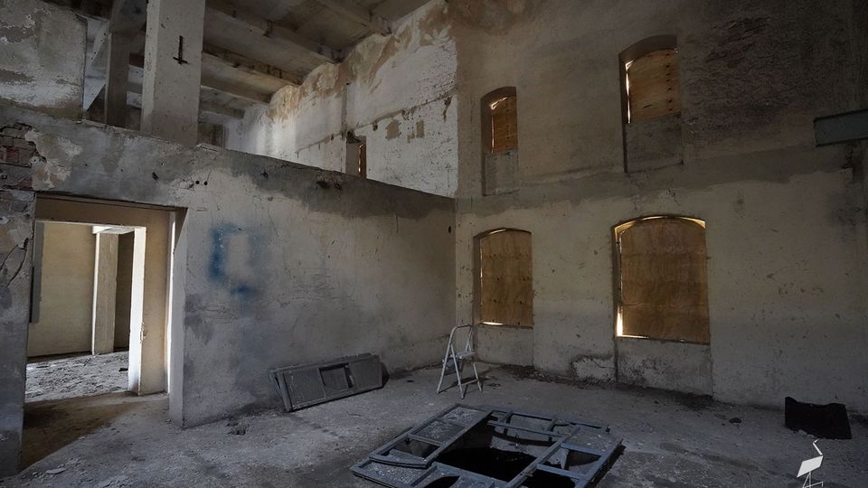 Innenraum eines verlassenen, zweigeschossigen Industrie- oder Lagergebäudes mit rohen Betonpfeilern, bröckelndem Putz und zugemauerten bzw. verbretterten Fensteröffnungen. Auf dem staubigen Boden liegen Metallrahmen und Schutt, in der Mitte befindet sich eine offene Bodenaussparung.