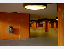 Unterirdischer Durchgang mit orangefarbenen Fliesenwänden und runden, beleuchteten Säulen. Ein Mülleimer ist an der linken Wand befestigt, und die Decke ist mit großen, runden Leuchten ausgestattet.