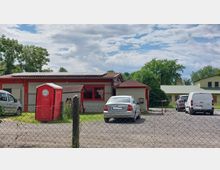 Ein einfacher Gebäudekomplex mit rotem und beigem Anstrich, davor ein Parkplatz mit mehreren geparkten Fahrzeugen und einer roten mobilen Toilette. Im Hintergrund sind grüne Bäume und ein weiteres Gebäude mit hellem Dach zu sehen, umgeben von einem Maschendrahtzaun.