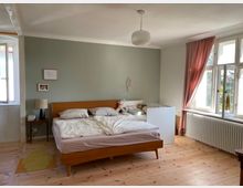 Schlafzimmer mit einem Doppelbett aus Holz, daneben ein weißes Kinderbett. Links steht ein kleiner Nachttisch mit Lampe, und rechts sind große Fenster mit rosa Vorhängen und einer darunterliegenden Heizung. Der Boden besteht aus hellem Holz, die Wand hinter dem Bett ist in einem grünlichen Farbton gestrichen.