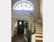 Heller Eingangsbereich eines Hauses mit geschwungener Holztreppe und verzierten weißen Geländern. Im Hintergrund eine braune Tür mit ovalen Fenstern und darüber ein großes, halbkreisförmiges Fenster mit farbiger Verzierung.