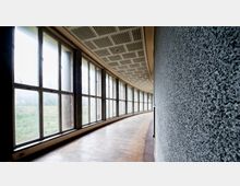 Gekrümmter Flur mit großflächigen Fensterfronten, durch die eine Wiese und Bäume sichtbar sind. Der Boden besteht aus Holz, die rechte Wand ist rau verputzt, und die Decke hat ein strukturiertes Muster.