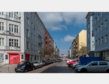 Straßenszene mit mehrstöckigen Wohngebäuden auf beiden Seiten, bemalt in neutralen und bunten Farben. Geparkte Autos säumen die gepflasterte Fahrbahn, während sich im Hintergrund ein modernes Gebäude mit einer Glasfront befindet.