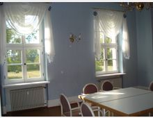 Ein heller Raum mit zwei großen Fenstern, verziert mit weißen, drapierten Vorhängen. Die Wände sind hellblau, es gibt zwei Heizkörper unter den Fenstern, sowie einen Tisch mit weißen Stühlen in der Mitte des Raumes.