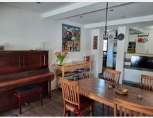 Esszimmer in einer Wohnküche mit Holztisch und Stühlen, einem Klavier an der Wand, einem Regal mit Büchern und Spielen sowie einer Vase mit roten Tulpen. Im Hintergrund eine offene Küche mit Kühlschrank, weißen Schränken und einer Theke, darüber eine Wanduhr.