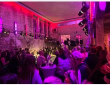 Ein Industriebau mit freiliegenden Ziegelwänden dient als Veranstaltungsort, beleuchtet durch farbiges, vorwiegend rosa Licht. Menschen stehen dicht gedrängt um eine beleuchtete Bühne, auf der eine Modenschau oder Darbietung stattfindet, während zahlreiche Scheinwerfer die Szene aus verschiedenen Winkeln erhellen.