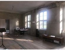 Ein spärlich möblierter Raum mit hohen Decken und großen Fenstern, die viel Tageslicht hereinlassen. Der Raum hat sichtbare, unverputzte Wände, einen Holzboden, einen runden schwarzen Tisch mit Stühlen, ein Regal mit Büchern und Dokumenten sowie ein Notenständer.