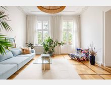 Helles Wohnzimmer mit hohen Decken, Stuckdetails und großem Fenster-/Balkontüren-Ensemble mit weißen Vorhängen. Parkettboden mit hellem Teppich, Glas-Couchtisch, Sofa, bunter Bodensessel sowie mehrere Zimmerpflanzen und eine große runde Deckenleuchte.