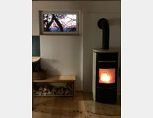 Ein schlicht eingerichteter Raum mit einem modernen Kaminofen mit brennendem Feuer auf der rechten Seite, darunter ein Glasplatten-Schutz auf Holzfußboden. Links steht eine Holzbank mit Feuerholz darunter und einem Korb darauf. Ein kleines Fenster im oberen Bereich zeigt Äste vor einem farbigen Abendhimmel.