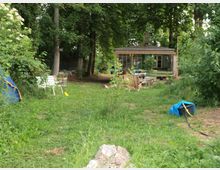 Waldlichtung mit einer kleinen Holzhütte im Hintergrund, umgeben von hohen Bäumen. Im Vordergrund befinden sich eine Zeltplane, Gartenmöbel und ein Lagerfeuerplatz, alles inmitten von Gras und Pflanzen.