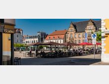 Städtischer Platz mit Cafés und roten Sonnenschirmen, gesäumt von historischen Gebäuden mit verzierten Fassaden und modernen Architekturstrukturen im Hintergrund. Mehrere Menschen sitzen an Tischen oder gehen in der sonnigen Umgebung.