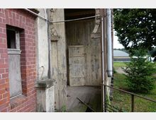 Teilansicht eines alten Gebäudes mit einer verwitterten Holztür und angedeutetem Holzvorbau. Links ist eine rote Backsteinmauer mit einem kleinen Fenster, rechts ein Metallgeländer, und im Hintergrund sind Bäume sowie ein landwirtschaftlich genutztes Gebäude zu sehen.