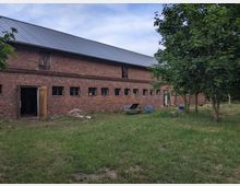 Eine lange, rechteckige Scheune aus rotem Ziegelstein mit kleinen rechteckigen Fenstern und einem grauen Blechdach, umgeben von einer Wiese mit einigen Bäumen und vereinzelten Gartenstühlen. Eine Holztür steht auf der linken Seite offen.