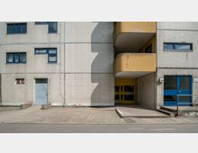 Wohngebäude in schlichter Bauweise mit heller, gefliester Fassade und blauen Fensterrahmen. Der Eingang ist gelb markiert und teilweise überdacht, während Balkone und Betonplattenwege den Außenbereich prägen.