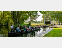 Eine Gruppe von Menschen fährt in einem traditionellen Holzkahn durch einen von Bäumen gesäumten Kanal. Rechts im Bild befindet sich ein Schleusentor aus Metall, daneben ein Holzsteg und eine Wiese mit Spaziergängern.