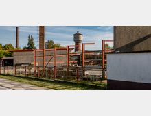 Industrielle Außenanlage mit teilweise errichteter Stahlkonstruktion vor einer Betonwand, im Hintergrund ein gemauerter Wasserturm und zwei hohe Schornsteine. Der Bereich ist durch einen Metallzaun abgegrenzt, umgeben von Bäumen und vereinzelten Gebäuden.