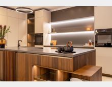 Moderne Wohnküche mit großer Kücheninsel aus Holz und grauer Arbeitsplatte, integriertem Spülbecken und Sitzbank. Helle, grifflose Schränke, eingebaute Backöfen, offene Regale mit indirekter LED-Beleuchtung und runde Deckenleuchte prägen den Raum.