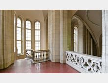 Innenansicht eines historischen Gebäudes mit hohen, schmalen Fenstern, durch die Tageslicht fällt. Der Raum hat massive Steinsäulen, eine verzierte Metallgeländer-Treppe und eine kunstvoll gestaltete Balustrade aus Stein.