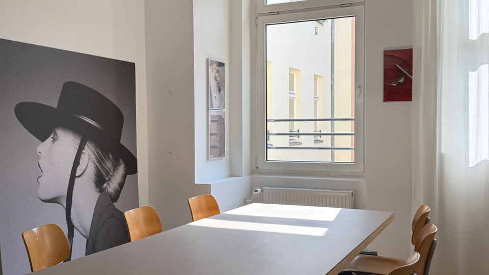Heller Besprechungsraum mit langem rechteckigem Tisch und mehreren Holzstühlen auf einem Teppich. Große Fenster mit weißen Vorhängen, runde Papier-Hängelampe und Wandkunst an den weißen Wänden.