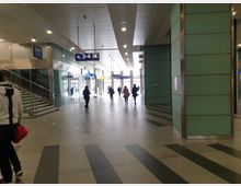 Innenansicht eines modernen Bahnhofs mit hohen Decken, grünen Wandverkleidungen und großen Glasflächen. Im Vordergrund ist eine Treppe sichtbar, im Hintergrund gehen Menschen durch einen breiten Gang in Richtung der Bahnsteige.