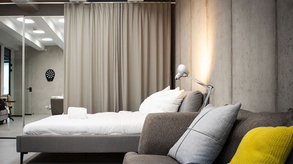 Modernes Schlafzimmer im Loft-Stil mit Betonwand, grauem Polsterbett mit weißer Bettwäsche und Stehleuchte neben dem Kopfteil. Im Vordergrund steht ein graues Sofa mit Kissen und gelber Decke; im Hintergrund trennt ein bodenlanger Vorhang den Raum, daneben sind Spiegelwand und Deckenbalken mit runden Leuchten zu sehen.