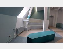 Minimalistisch gestalteter Innenraum mit pastellfarbenen Wänden in Blau und Weiß. Auffällig sind schräge Wände, eine Sitzfläche mit Holzdetails, ein Heizkörper und geometrische Elemente, darunter ein türkisfarbener Tisch.