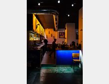 Eine Bar mit dimmiger Beleuchtung; links beleuchtete Regale mit Flaschen und ein Tresen, im Hintergrund Dekorationen wie Tierköpfe an der Wand und farbige Kunstwerke. Rechts im Vordergrund eine blaue Barfront mit einem einzelnen Holzstuhl davor.