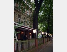 Außenansicht eines Cafés oder Restaurants mit überdachter Terrasse, umgeben von einem Zaun aus Holz und Backsteinpfeilern, flankiert von großen Bäumen. Laternen entlang des Gehwegs sorgen für Beleuchtung, und ein Mann arbeitet an einem Tresen im vorderen Bereich des Gebäudes.