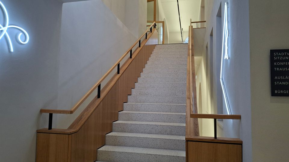 Modernes Treppenhaus in einem öffentlichen Gebäude mit breiter, hellgrauer Steintreppe und Holzgeländern an beiden Seiten. Weiße Wände mit eingelassenen Fensteröffnungen und leuchtenden Wandinstallationen sowie einem Richtungsschild entlang des Treppenaufgangs.
