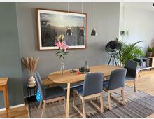 Esszimmer mit einem Holztisch und fünf grauen Polsterstühlen auf einem Teppich. An der Wand hängt ein großes gerahmtes Strandfoto, daneben befinden sich Pflanzen und eine Stehlampe; Dekorationen wie eine Vase mit Blumen und Kerzen stehen auf dem Tisch.