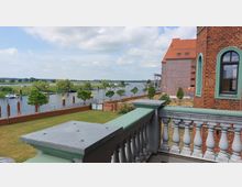 Blick von einem Balkon mit weißem Geländer und grünen Akzenten auf einen Fluss mit vertäuten Booten, umgeben von grünen Wiesen und Bäumen. Im Hintergrund sind Backsteingebäude mit roten Dächern sowie eine flache, ländliche Landschaft zu sehen.