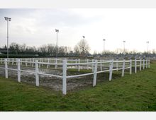 Eine umzäunte Außenfläche mit weißem Gatter auf schlammigem Boden, umgeben von einer Wiese. Im Hintergrund befinden sich hohe Flutlichtmasten, Bäume und Gebäude am Horizont.