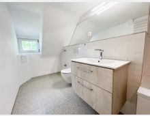 Modernes Badezimmer mit einer Dachschräge, einem Waschbecken mit Holzunterschrank, einer großen eingelassenen Spiegelwand und einem integrierten Wand-WC. Der Boden ist mit gemusterten Fliesen und die Wände teils mit weißen Kacheln verkleidet; ein kleines Fenster bietet Tageslicht.