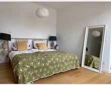 Schlafzimmer mit einem Doppelbett, das eine grüne Bettdecke mit Blumenmuster hat. Auf beiden Seiten des Bettes stehen Holznachttische mit dekorativen Lampen, und ein großer, weiß gerahmter Spiegel lehnt an der rechten Wand.