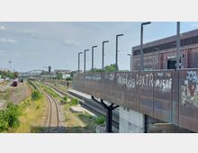 Blick auf den Bahnhof Warschauer Straße in Berlin mit Gleisanlagen im Vordergrund, umgeben von Bahngleisen und Vegetation. Rechts ist ein modernes Gebäude mit Graffiti an der Fassade und einer ausgeschilderten S-Bahn-Station sichtbar, im Hintergrund ein Bogenbrücke und ein Wasserturm.