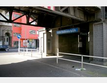 U-Bahnhof Nollendorfplatz in Berlin, aufgenommen unter einem Eisenbahnviadukt mit sichtbarer Stahlkonstruktion. Im Hintergrund befinden sich eine orangefarbene Gebäudefassade, Straßenschilder und ein Gehweg.