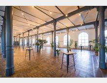 Großer, heller Veranstaltungsraum mit hohen Decken, säulenartiger Stahlkonstruktion, Parkettboden und großen halbkreisförmigen Fenstern. Der Raum ist mit vereinzelten Stehtischen, Pflanzen in Töpfen und Lichterketten dekoriert.