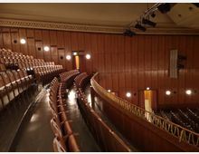 Ein Theater- oder Konzertsaal mit hölzerner Wandverkleidung und Reihen brauner Sitzplätze, die sich in einem halbkreisförmigen Aufbau erstrecken. An der Decke befinden sich Beleuchtungs- und Bühnentechnik, und Türen mit Vorhängen führen in angrenzende Bereiche.