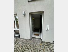 Eingangsbereich eines Gebäudes mit einer offenen Tür, die in einen kleinen Flur mit Fliesenboden und Fußmatten führt. Vor der Tür ist ein gepflasterter Boden mit einem Metallgitter zu sehen, daneben eine Gegensprechanlage an der Wand.