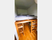 Holzhochbett in einem kleinen Zimmer mit weiß gestrichenen Wänden. Unter dem Hochbett befinden sich ein Regal und eine Leiter, der Boden besteht aus hellem Parkett.